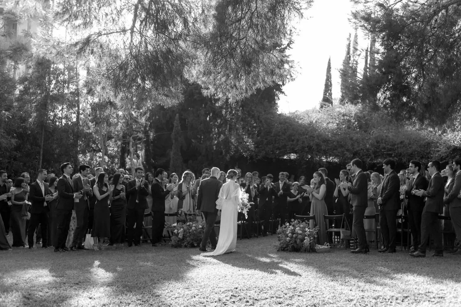 A black and white photo of a bride and groom walking hand-in-hand through a row of guests, who are standing and clapping in celebration right after the couple has exchanged vows. The moment captures a joyful, intimate atmosphere as the newlyweds move through the crowd, surrounded by the support and love of their guests.